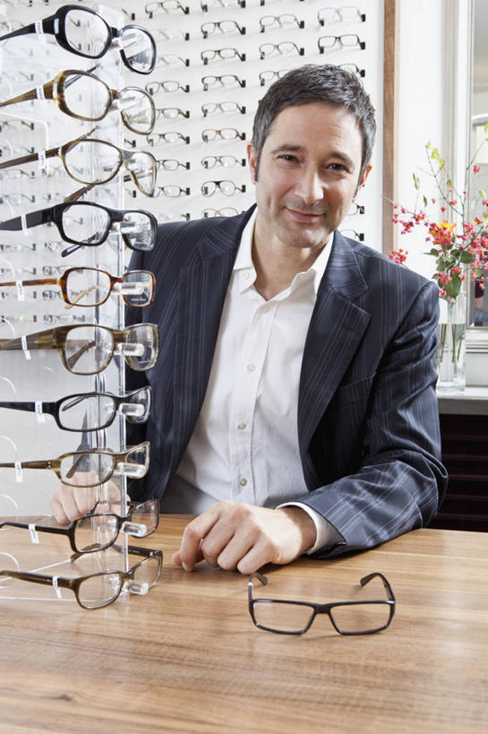 西方人,一个人,商店,坐,笑,微笑,露齿笑,竖图,室内,白天,正面,西装,桌面,桌子,欧洲,欧洲人,仅一个男性,仅一个人,仅一个中年男性,套装,正装,朦胧,模糊,木制,眼镜,注视,职业装,商铺,货架,架子,木制品,观察,看,观看,验光师,察看,关注,陈列架,近视眼镜,眼镜店,超市货架,市肆,商号,男人,男性,中年男性,白种人,坐着,半身,彩图,店铺