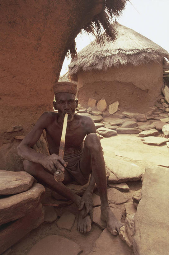 西方人,成年人,老年人,皱纹,胡须,一个人,家,坐,吸烟,竖图,室外,白天,正面,旅游,度假,美景,帽子,欧洲,非洲,贝宁,黑人,欧洲人,仅一个男性,仅一个人,房屋,屋顶,屋子,草屋,景观,泥土,乡村,陶瓷制品,图像,顶棚,拿着,娱乐,住宅,衣服,天空,自然,茅草屋,看,仅一个老年男性,享受,休闲,赤脚,躯干,鸭舌帽,陶器,景色,放松,人体,摄影,泥,毛发,住房,贫穷,自然风光,主题,自然现象,视角,构图,西非,萨满,老年男性,男人,男性,非洲人,胸部,胡子,络腮胡,小胡子,岩石,彩图,旅行,房子,茅舍,茅屋,小屋,时空,传统文化,发展中国家,社会问题